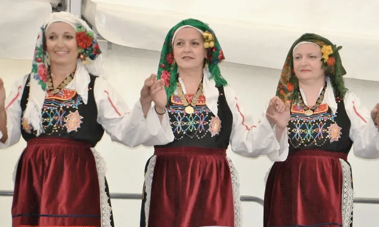 Drei Frauen in traditioneller Kleidung stehen und klatschen. Sie tragen rote und weiße Kleider mit Blumenkopfbedeckungen.