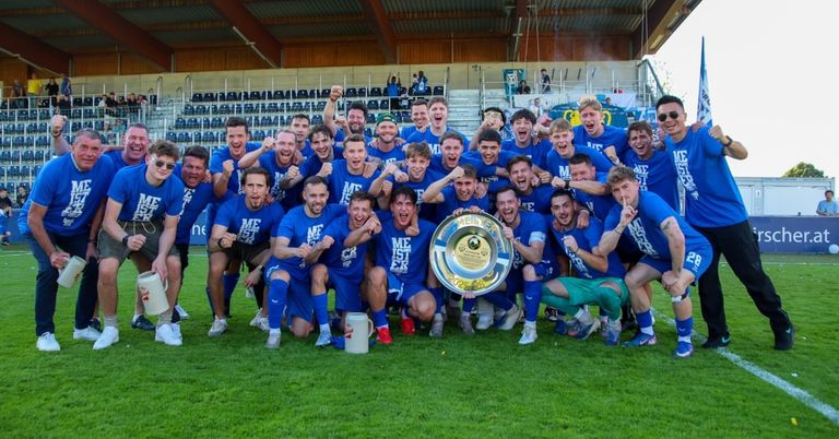 Eine Fußballmannschaft feiert mit einem Pokal auf dem Feld, mit blauen Trikots und Fans im Hintergrund.