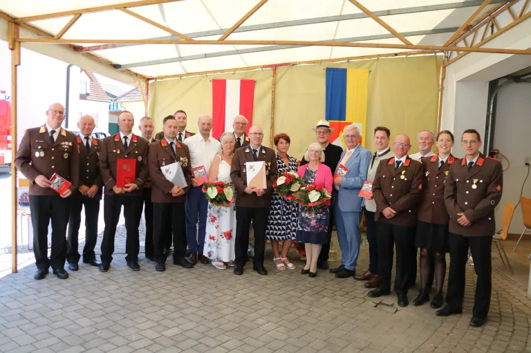 Eine Gruppe von Menschen in Uniform und ziviler Kleidung steht unter einem Zelt mit Fahnen. Sie halten Auszeichnungen und Blumen.