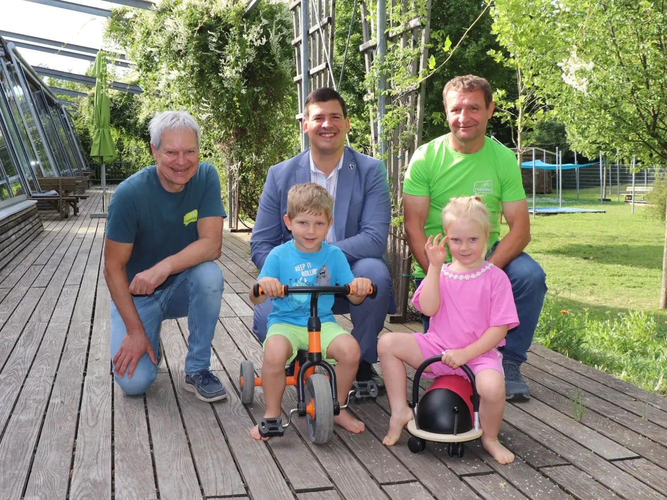 Drei Männer und zwei Kinder, ein Mädchen und ein Junge, posieren für ein Foto auf einer Holzterrasse. Der Junge fährt ein Dreirad, während das Mädchen auf einem Spielzeug sitzt.