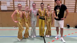 Eine Gruppe junger Mädchen in Goldkostümen mit Medaillen um den Hals posiert mit einer Trophäe in einer Turnhalle für ein Foto.
