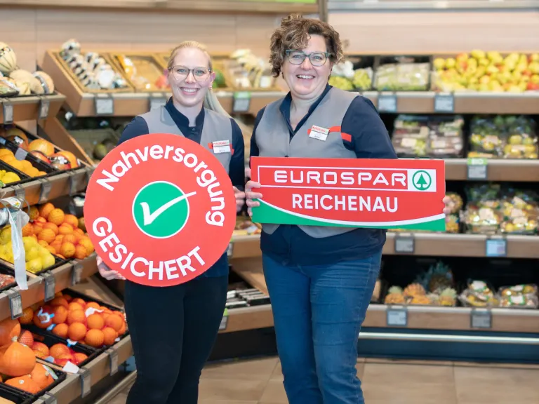 Zwei Frauen in grauen Westen stehen in einem Supermarkt, lächeln und halten Schilder. Das linke Schild sagt 'Nahversorgung gesichert' und das rechte Schild sagt 'EUROSPAR REICHENALU'. Sie stehen vor Regalen mit Obst und Gemüse.