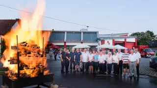 Eine Gruppe von Menschen steht vor einem Feuerkorb mit einem brennenden Feuer und einem Gebäude mit dem Schild Feuerwehrhaus im Hintergrund.