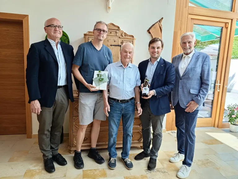 Fünf Männer stehen in einer Reihe und lächeln. Der zentrale Mann hält eine Karte mit einer Pflanze. Der Mann zu seiner rechten hält eine Flasche. Dahinter steht ein hölzerner Schrank an einer Wand.