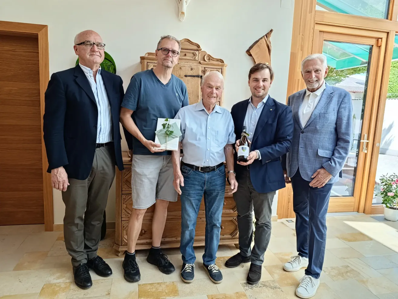 Fünf Männer stehen in einer Reihe und lächeln. Der zentrale Mann hält eine Karte mit einer Pflanze. Der Mann zu seiner rechten hält eine Flasche. Dahinter steht ein hölzerner Schrank an einer Wand.