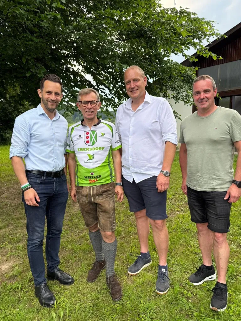 Four men are standing in a field, smiling. The man in the center wears a green and white shirt with the word 'Ebersdorf' on it. The man on the left wears a light blue shirt. The man on the right wears a green shirt. The man in the middle wears a white shirt and dark shorts.