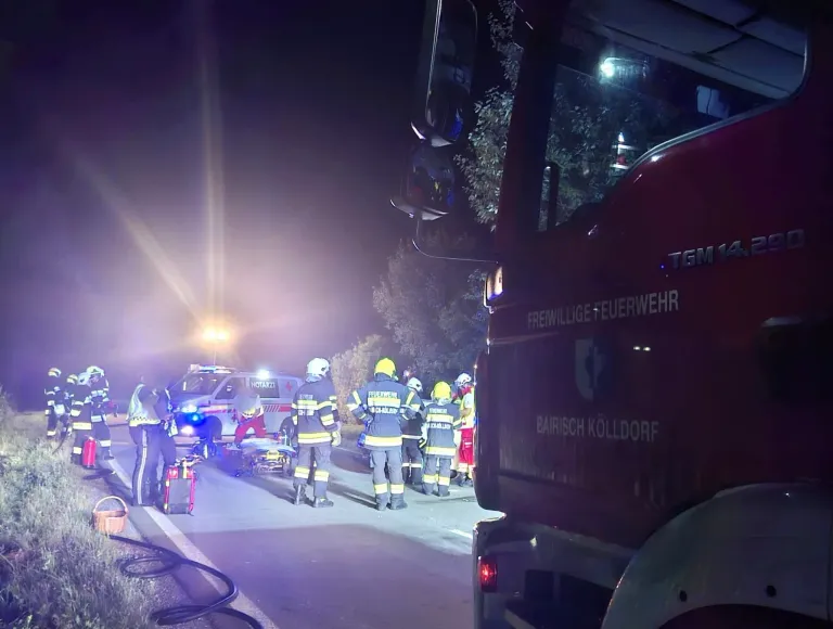 Feuerwehrleute und Sanitäter bereiten sich darauf vor, eine verletzte Person auf einer Trage zu transportieren. Die Szene wird von den Lichtern der Rettungsfahrzeuge beleuchtet.
