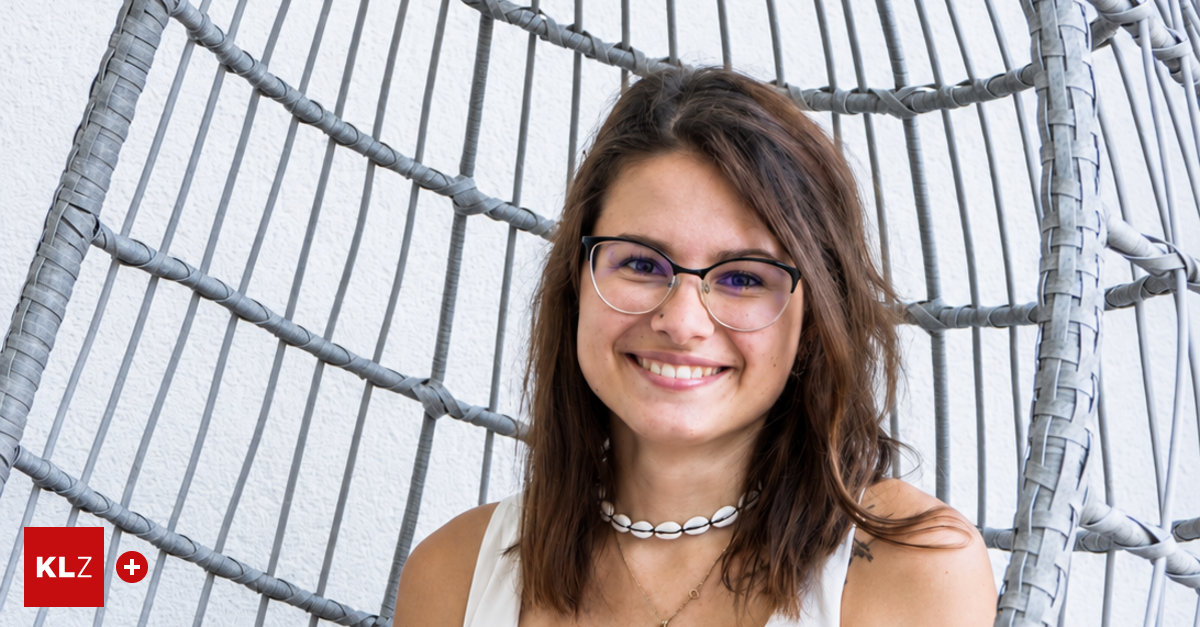 Eine Frau mit braunen Haaren und einer Brille lächelt in die Kamera. Sie trägt ein weißes ärmelloses Top und eine weiße Muschelkette. Sie befindet sich in einem Raum mit grauem Metallhintergrund.