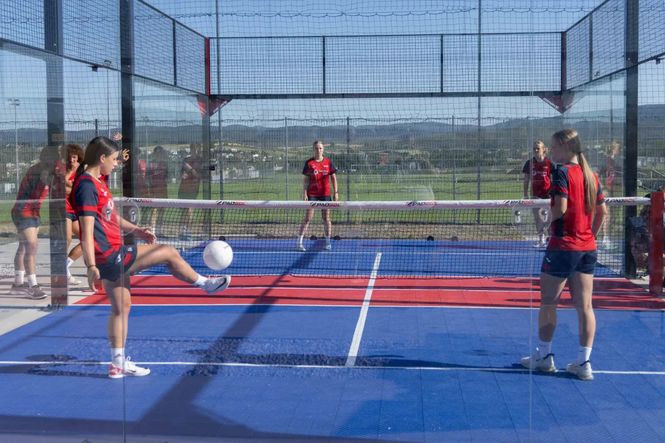 Eine Person in Rot tritt einen Ball auf einem blauen Platz mit Netz, während andere in Rot zusehen. Ein Zaun und Berge sind im Hintergrund.