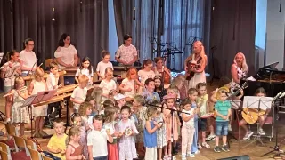 Eine Gruppe von Kindern in verschiedenen farbigen Outfits steht mit Musikinstrumenten auf einer Bühne, begleitet von einigen Erwachsenen, und sie scheinen an einer musikalischen Aufführung teilzunehmen.