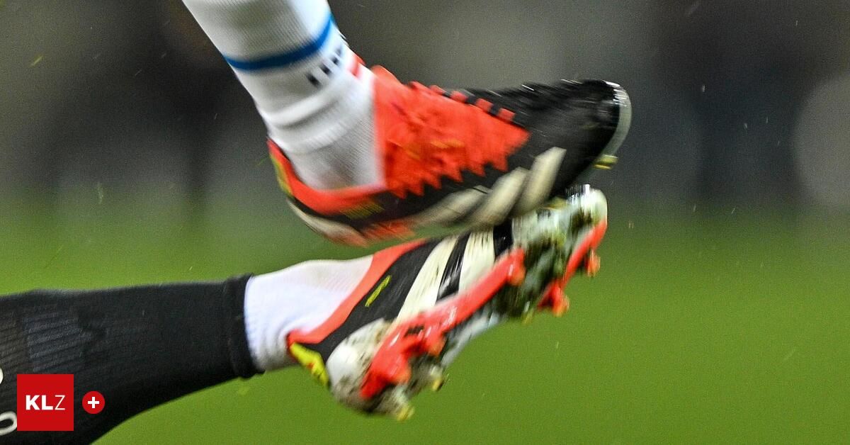 Fußballspieler mit roten und schwarzen Stollenschuhen und weißen Socken, der den Ball tritt, in Bewegung, auf einem grünen Feld.