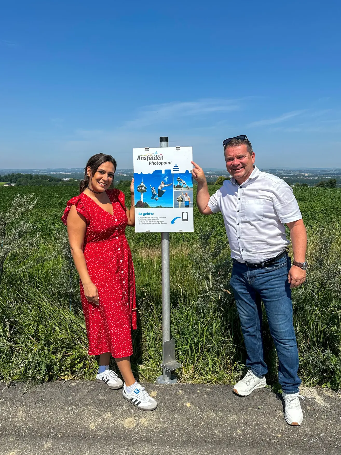 Ein Mann und eine Frau posieren neben einem Fotopunkt-Schild auf einem Feld. Die Frau trägt ein rotes Kleid und Turnschuhe. Der Mann trägt ein weißes Hemd, Jeans und Turnschuhe.