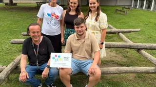 Eine Gruppe von Menschen posiert für ein Foto mit einem Schild für Mint Gutesiegel 2026 vor sich. Der Mann auf der linken Seite hält ein Zertifikat, während die anderen lächeln. Dahinter sind ein Holzzaun und eine grasbewachsene Fläche zu sehen.