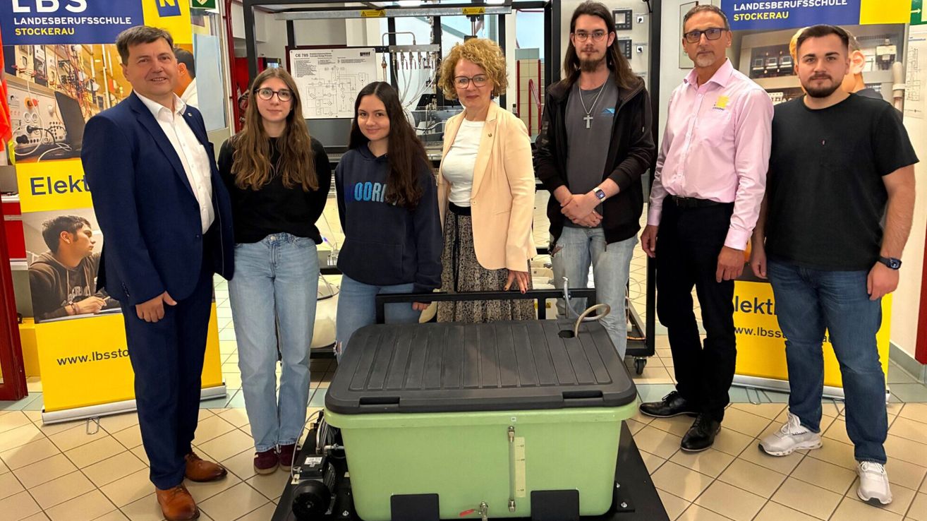 Fünf Personen stehen um eine grüne Aufbewahrungsbox. Zwei Frauen, zwei Männer und eine Frau in einer weißen Jacke. Die Frauen tragen eine Brille und Jeans. Dahinter ist eine Tafel mit technischen Zeichnungen an der Wand befestigt.
