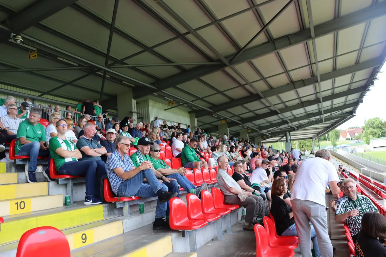 Ein Stadion voller Zuschauer, die Grün und Weiß tragen, einige sitzen auf roten Stühlen, andere stehen. Die Atmosphäre ist lebhaft, mit Leuten, die sich unterhalten und die Veranstaltung genießen.