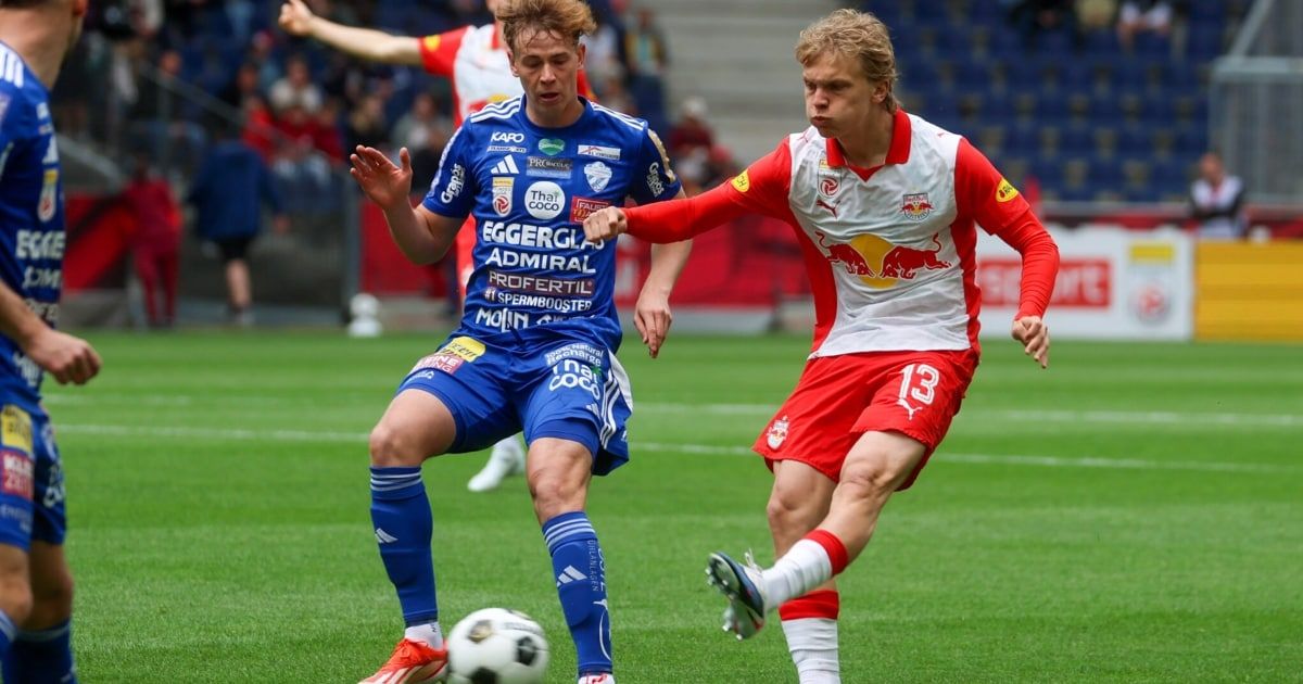 Zwei Fußballspieler in blauen und roten Trikots sind auf einem Feld. Ein Spieler tritt den Ball, während der andere verteidigt.