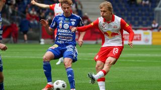 Zwei Fußballspieler in blauen und roten Trikots sind auf einem Feld. Ein Spieler tritt den Ball, während der andere verteidigt.