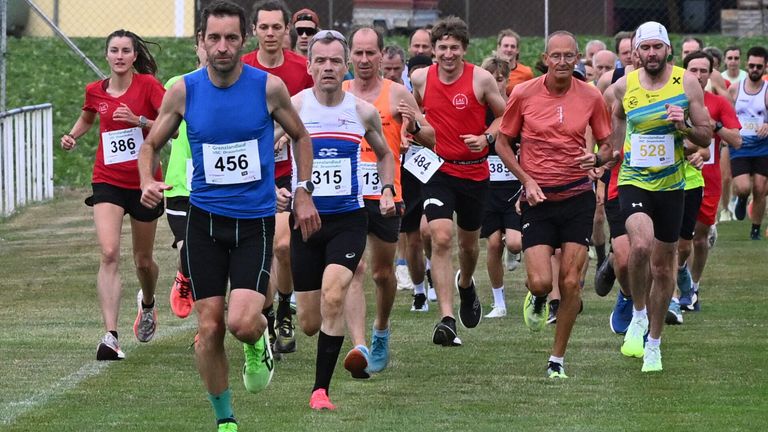 Several runners compete in a race. One runner wears a blue shirt with the number 456. Another wears a red shirt with the number 389.