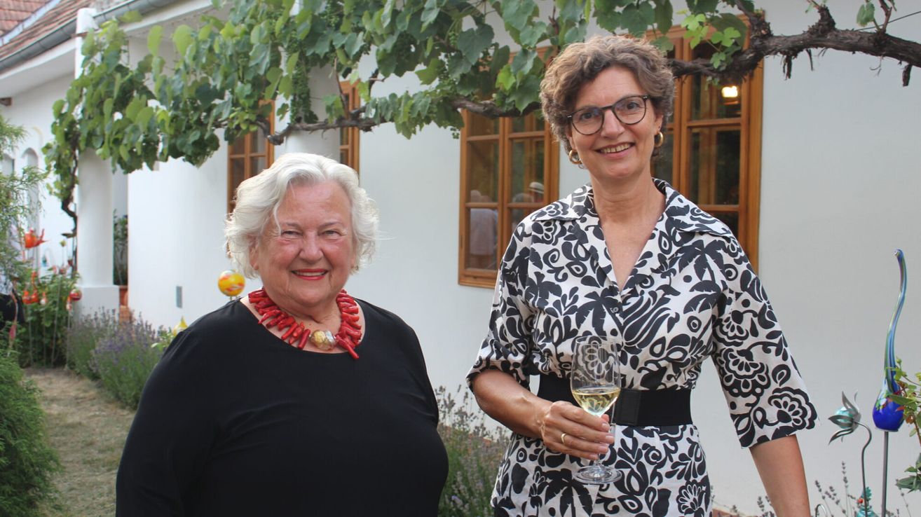 Two women stand outside a building. One woman holds a wine glass, the other wears a red necklace. Grapes hang from a tree above them.