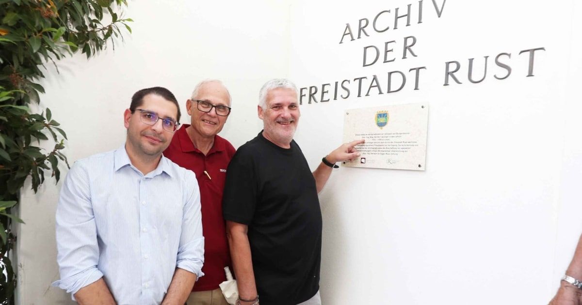 Three men stand next to a plaque mounted on a white wall. The man in the middle points at the plaque.