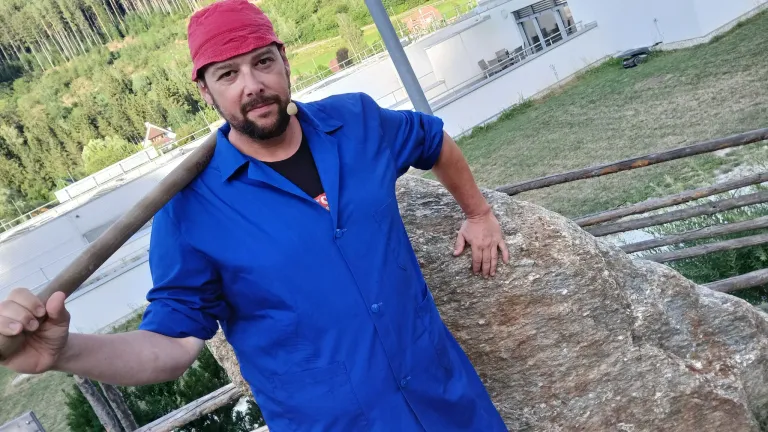 A man in a blue shirt and red cap holds a wooden bat while standing next to a large rock and a white building.