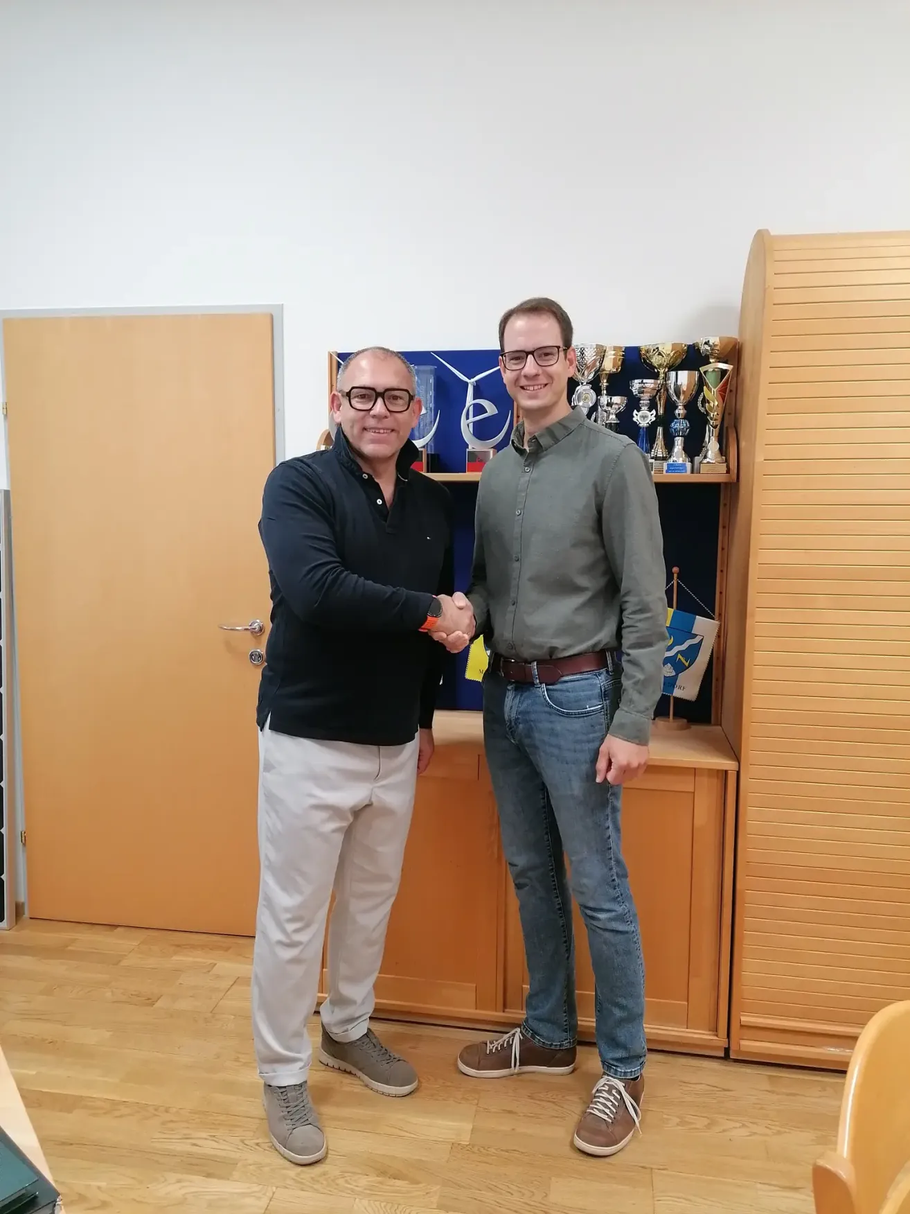 Zwei Männer schütteln sich die Hände vor einem Schrank mit Trophäen und einer Flagge. Sie tragen beide eine Brille und lächeln.