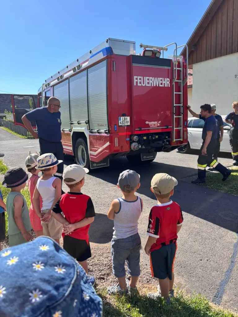 A group of children, some in hats, watch a firefighter explaining something about a red fire truck with the word 'Feuerwehr' on it.