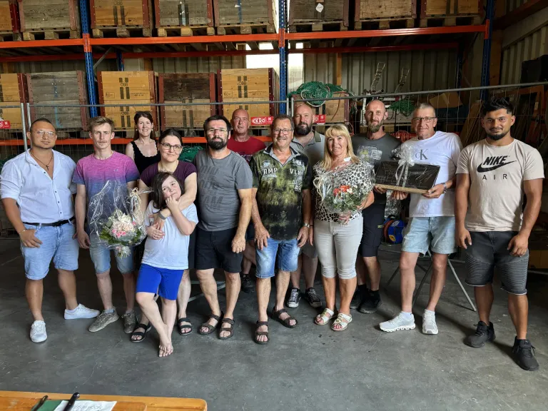 Eine Gruppe von Menschen posiert für ein Foto in einem Lagerhaus mit Regalen und Kisten im Hintergrund. Sie halten Blumen und eine Plakette und stehen in lässiger Kleidung.