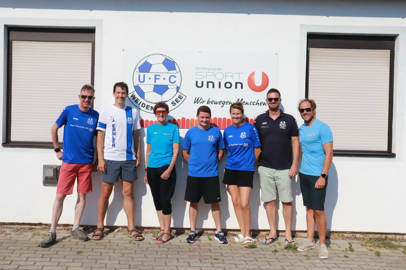 Seven people are posing for a photo in front of a white wall with a logo. The group includes both men and women, all wearing light blue shirts. They are standing in a row, smiling at the camera.