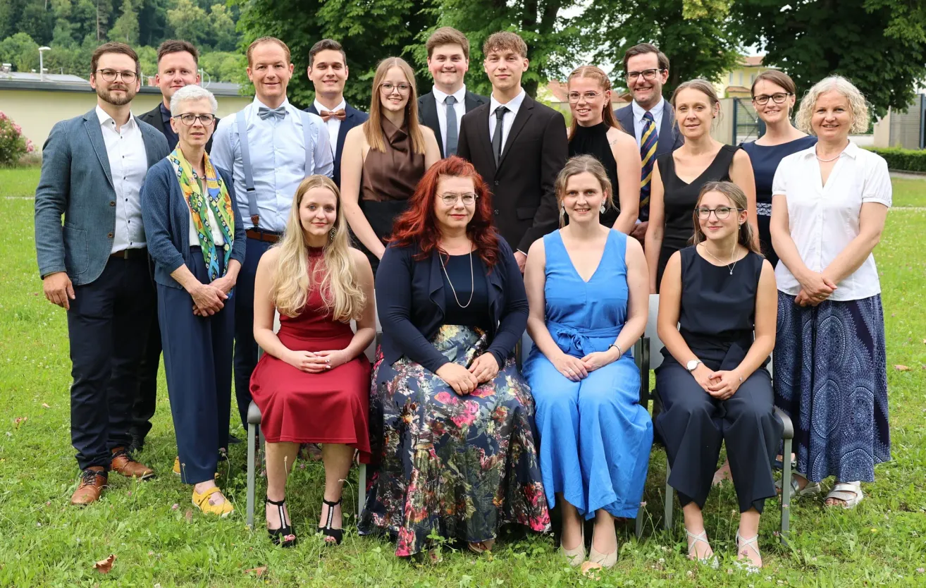 Eine Gruppe von Menschen in formeller Kleidung posiert für ein Foto auf einer Rasenfläche mit Bäumen im Hintergrund. Einige sitzen, während andere hinter ihnen stehen.