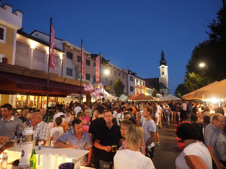 At night, a crowded street is filled with people and tents. There are flags and a tower in the background. Many people are standing and sitting, some with drinks. The buildings are lit up, and the sky is clear.