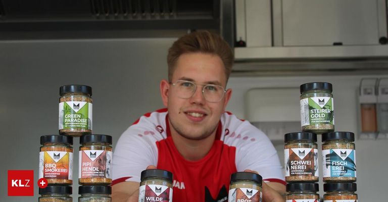 A man wearing a red and white shirt stands in a kitchen, smiling, with various jars of spices behind him. The jars have labels like 'Wilde' and 'Brot'. He holds a jar in his right hand.