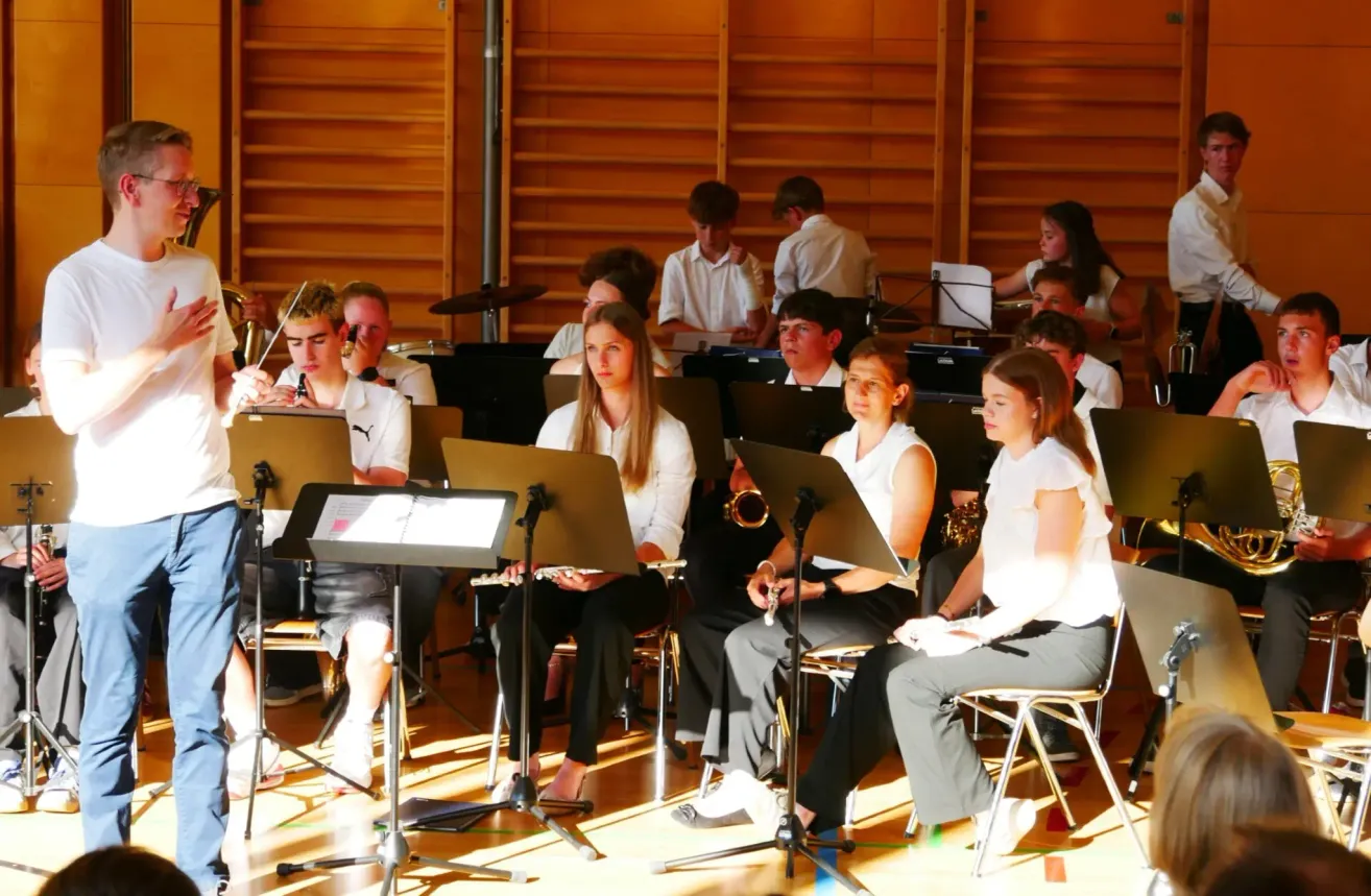 A group of students in white shirts and black pants are playing instruments like the flute and trumpet in a room with wooden shelves.