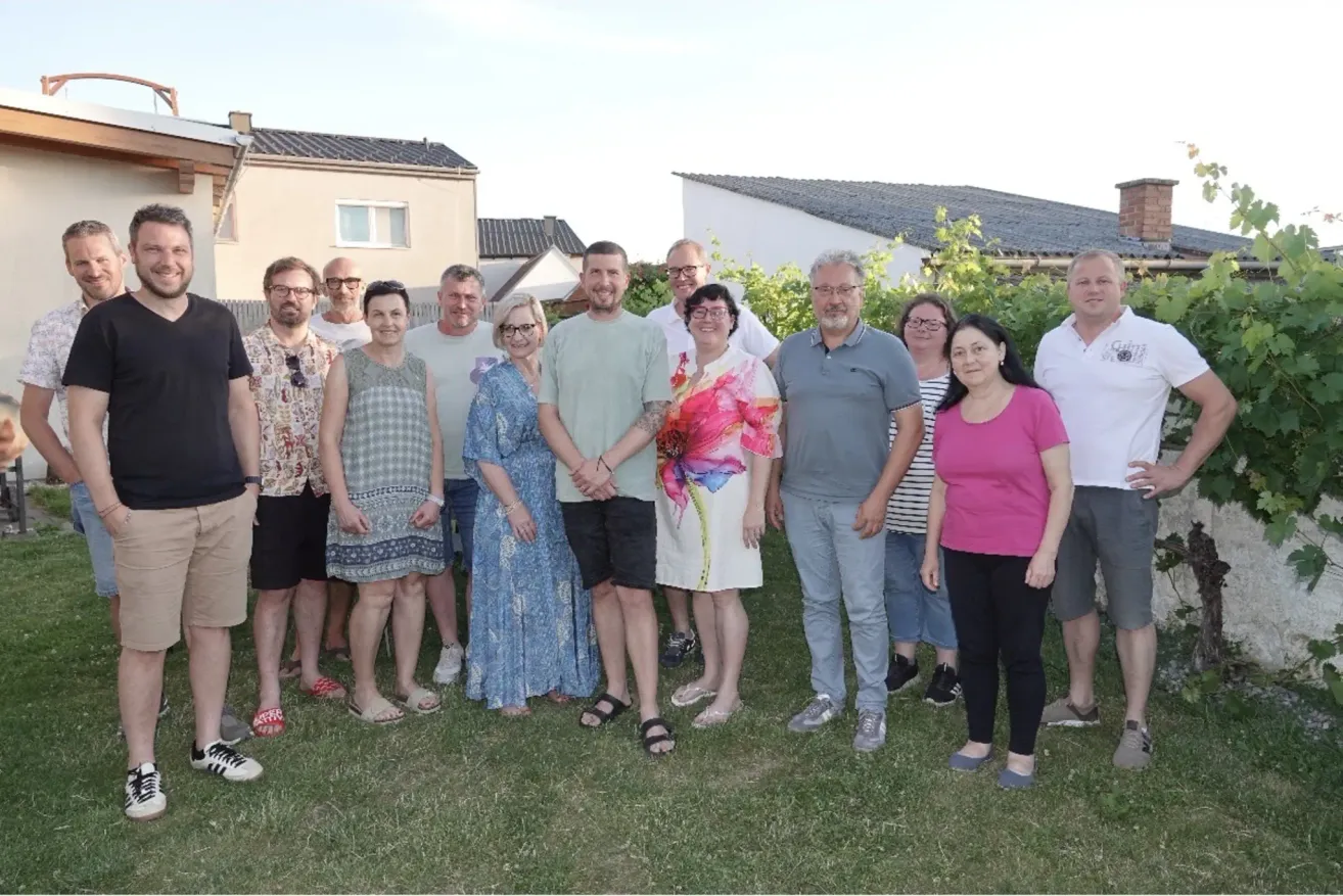 Eine Gruppe von Männern und Frauen steht auf einem Rasen und posiert möglicherweise für ein Foto. Sie tragen lässige Kleidung. Im Hintergrund befinden sich Gebäude.