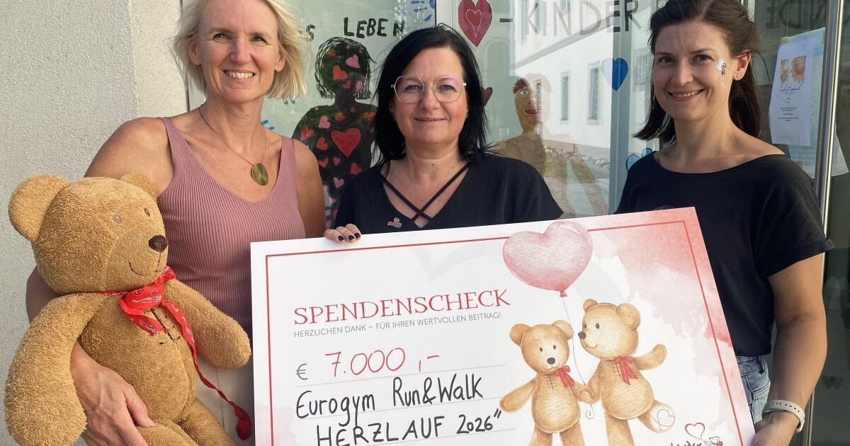 Drei Frauen stehen lächelnd vor einem Schaufenster. Die Frau in der Mitte hält einen Spendenscheck über 7.000 Euro. Die Frau auf der linken Seite hält einen Teddybären und die Frau auf der rechten Seite hält einen herzförmigen Ballon. Das Schaufenster zeigt Zeichnungen und Herzen.