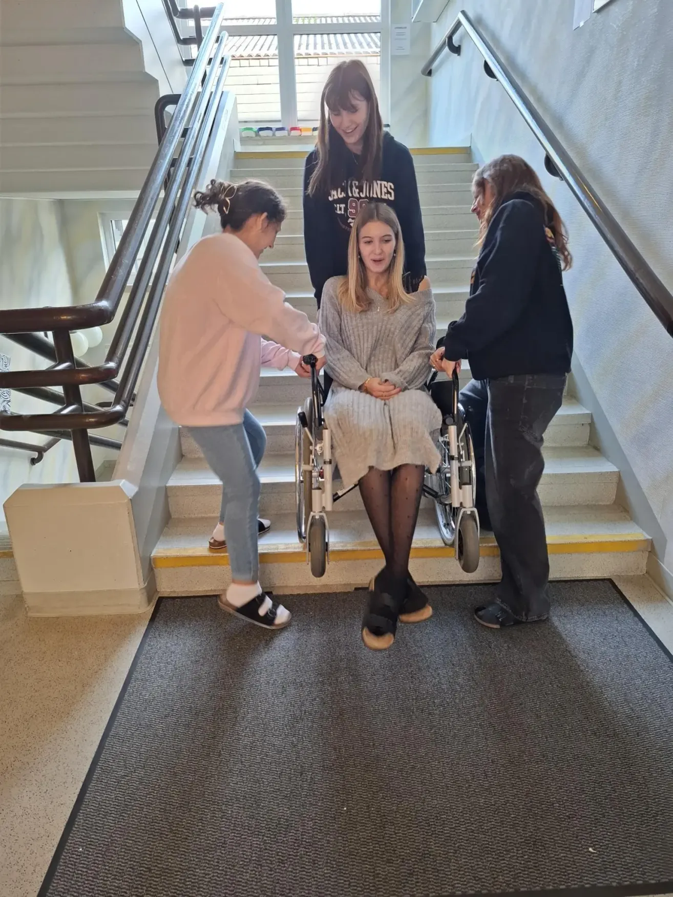 Three women assist a girl in a wheelchair on a flight of stairs. The girl smiles.