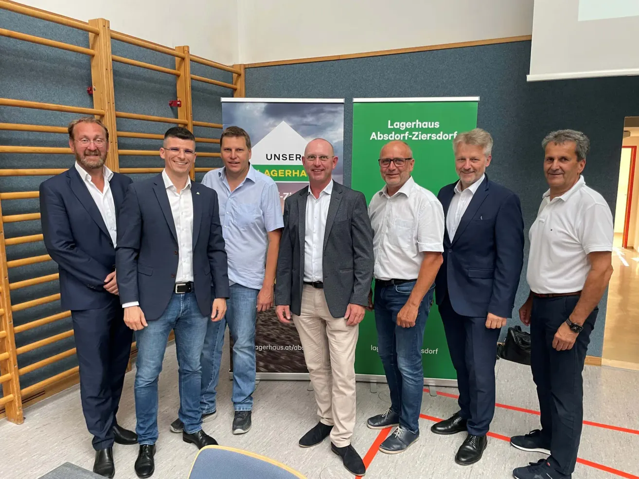 Six men stand together in front of a banner that reads Lagerhaus Absorf-Ziersdof. They are all dressed in suits and smiling for the camera.