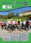 Eine Gruppe von Menschen steht mit Fahrrädern vor einem grünen Banner. Ein Mann sitzt auf dem Boden mit einem Fahrrad vor ihm. Ein Tisch steht hinter ihm. Dahinter befindet sich ein Haus mit einem Baum davor.