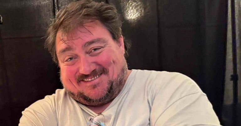 A man with a beard and a white t-shirt smiles and poses for a photo. He has glasses in his hand.