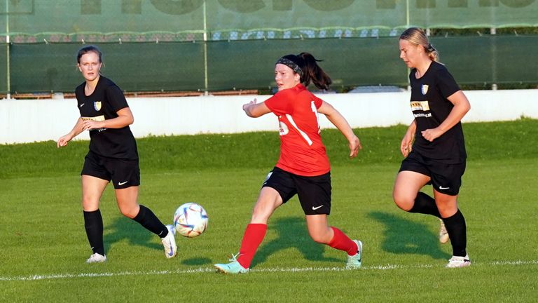 Three women are playing soccer. One player in a red jersey is running towards the ball. Another player in black runs alongside. The third player, also in black, is looking towards the ball.