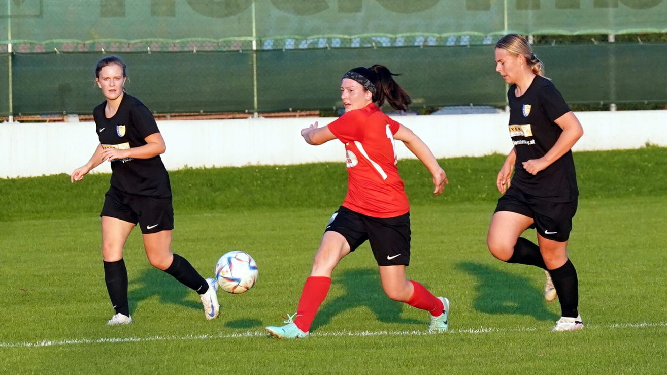 Three women are playing soccer. One player in a red jersey is running towards the ball. Another player in black runs alongside. The third player, also in black, is looking towards the ball.