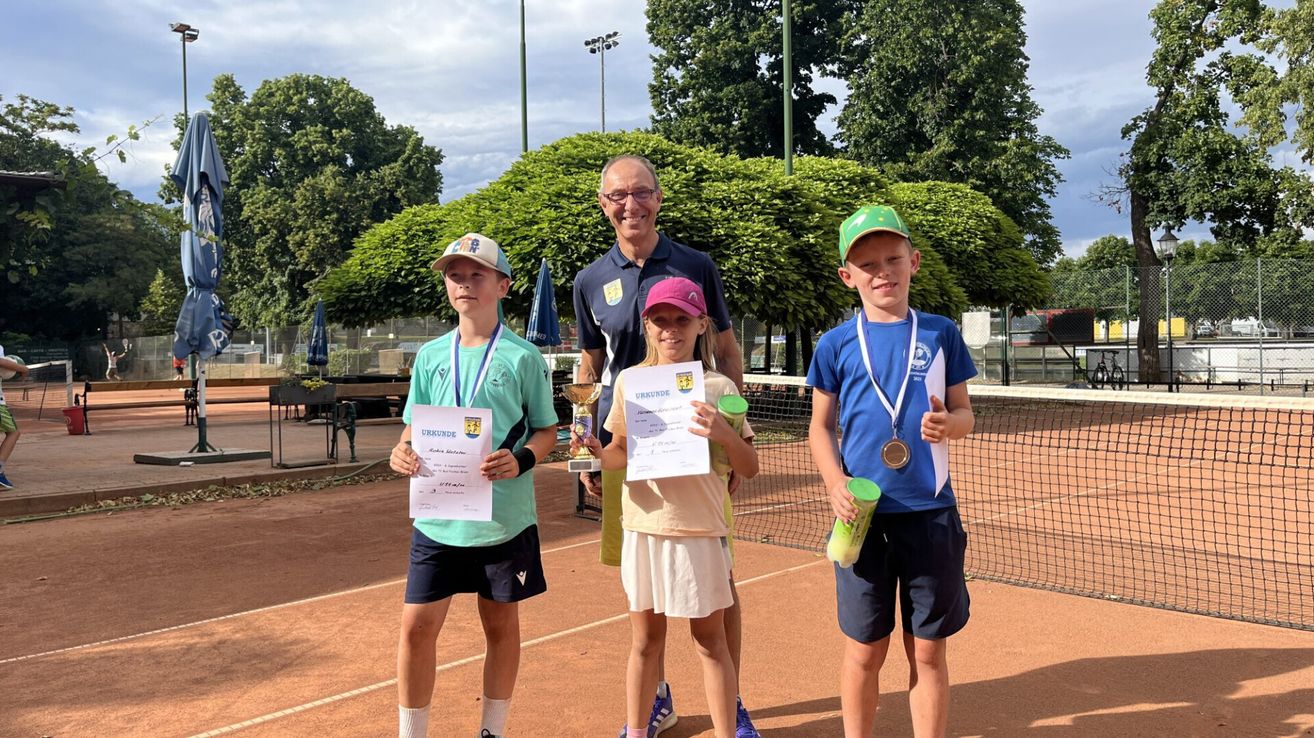 Three kids stand on a tennis court with medals and certificates, smiling for a photo. Behind them is a man wearing a polo shirt and standing next to a hedge.
