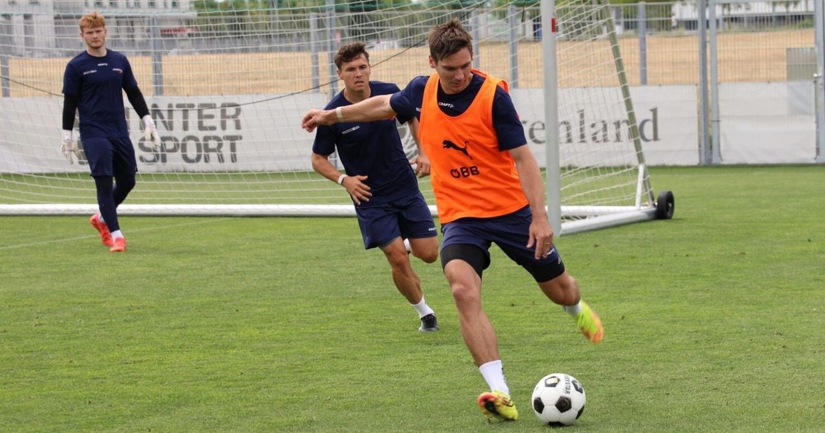 Two men play soccer on a field with a goal. One man in an orange vest is kicking the ball. The other man is running towards him.