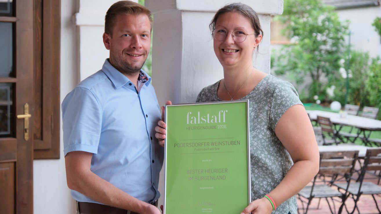 Ein Mann und eine Frau halten ein gerahmtes Falstaff-Auszeichnungszertifikat. Der Mann trägt ein blaues Hemd und die Frau trägt ein Blumenhemd und eine Brille.