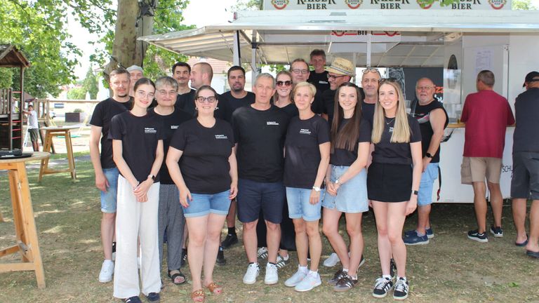 A group of people in matching black t-shirts stands in front of a Kaiser Bier bar cart. The setting is outdoors with a canopy overhead and trees in the background.