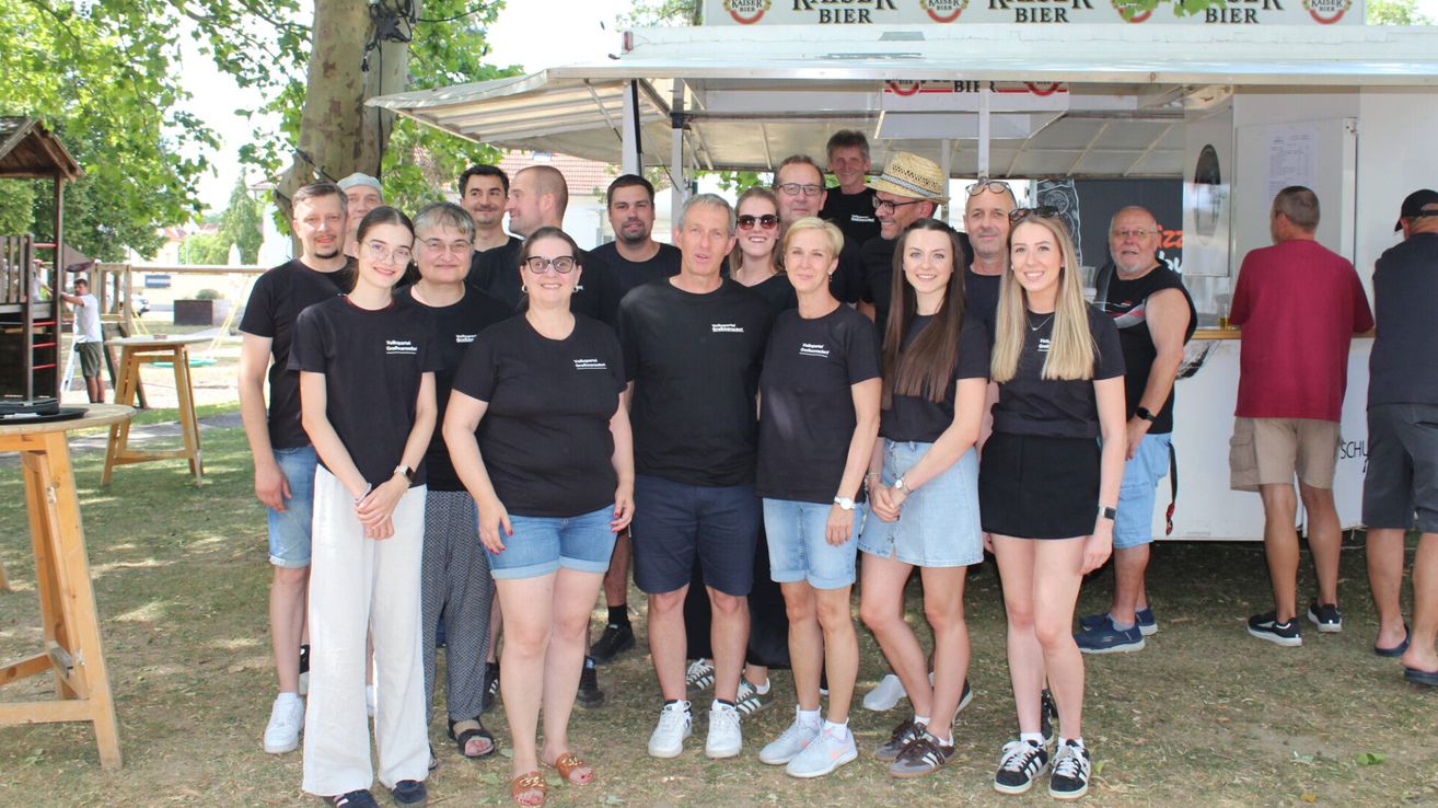 Eine Gruppe von Menschen in passenden schwarzen T-Shirts steht vor einem Kaiser Bier Barwagen. Die Umgebung ist im Freien mit einem Vordach und Bäumen im Hintergrund.