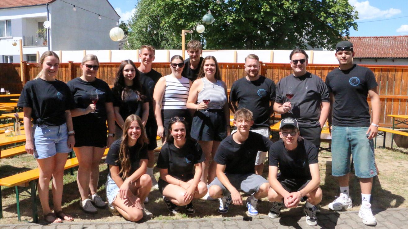 A group of people in matching black shirts pose for a photo. They are holding wine glasses. Behind them is a wooden fence and some trees.