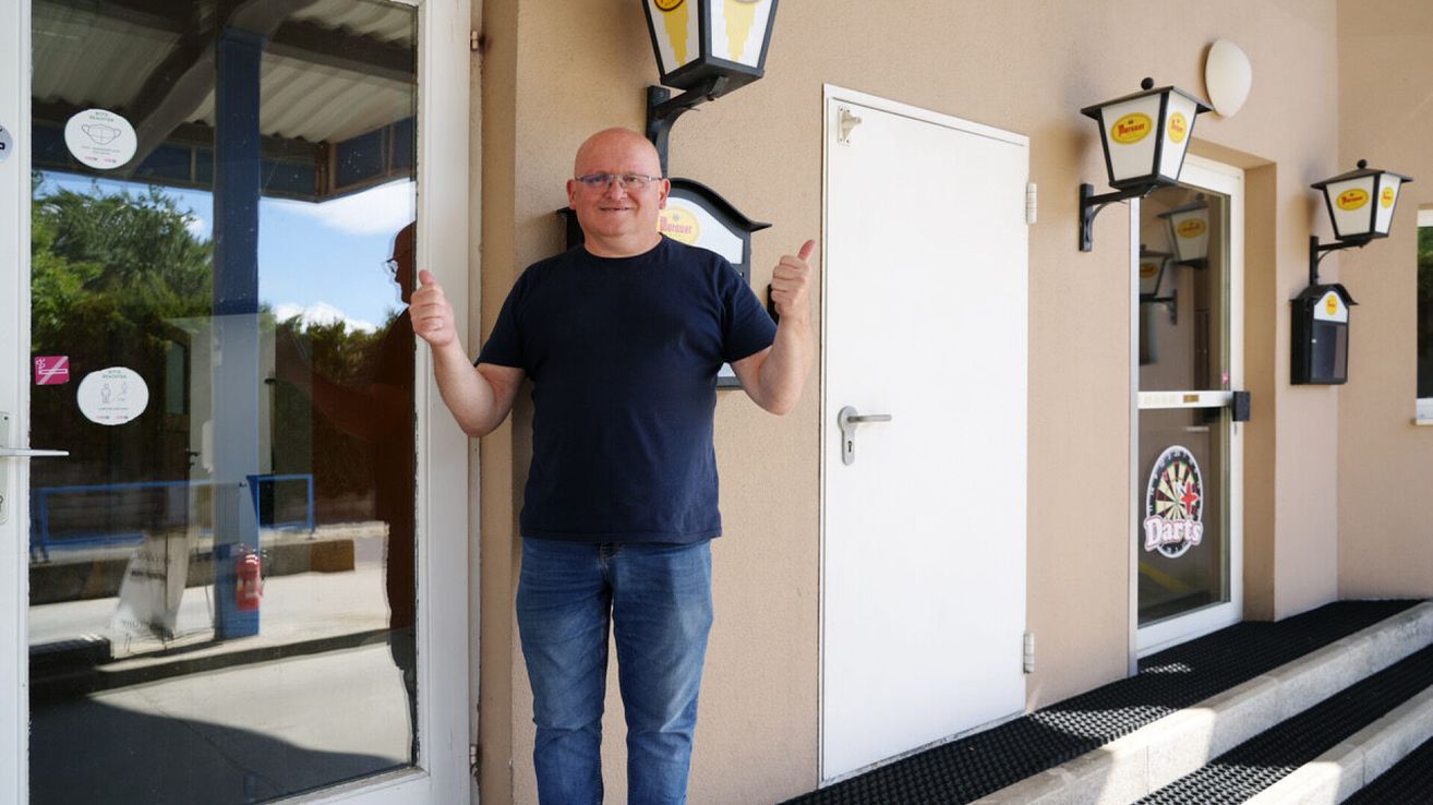 A man in a blue shirt and jeans gives a thumbs up while standing in front of a building. There is a lamp on the wall and a door nearby.