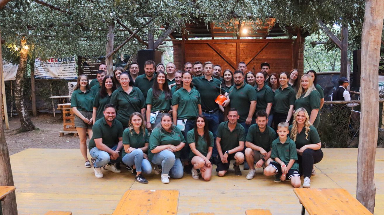 A group of people in green uniforms, possibly employees, pose for a photo in an outdoor area with a wooden structure and trees.