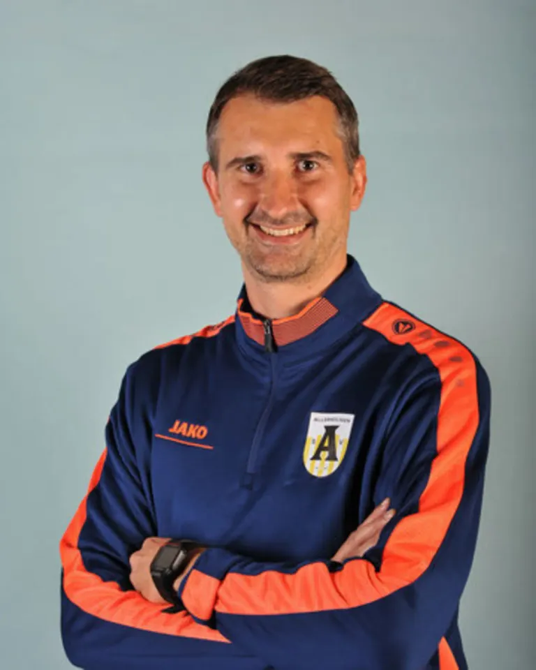 A man smiles while standing with folded arms, wearing a blue and orange sports jacket with 'Jako' and 'A' logos. He appears to be a sports professional.
